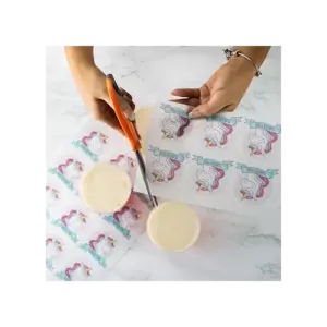 Hands using scissors to cut edible cake toppers featuring unicorn designs on a flat surface.