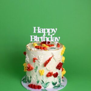 A birthday cake decorated with flowers and a 'Happy Birthday' topper on a green background.