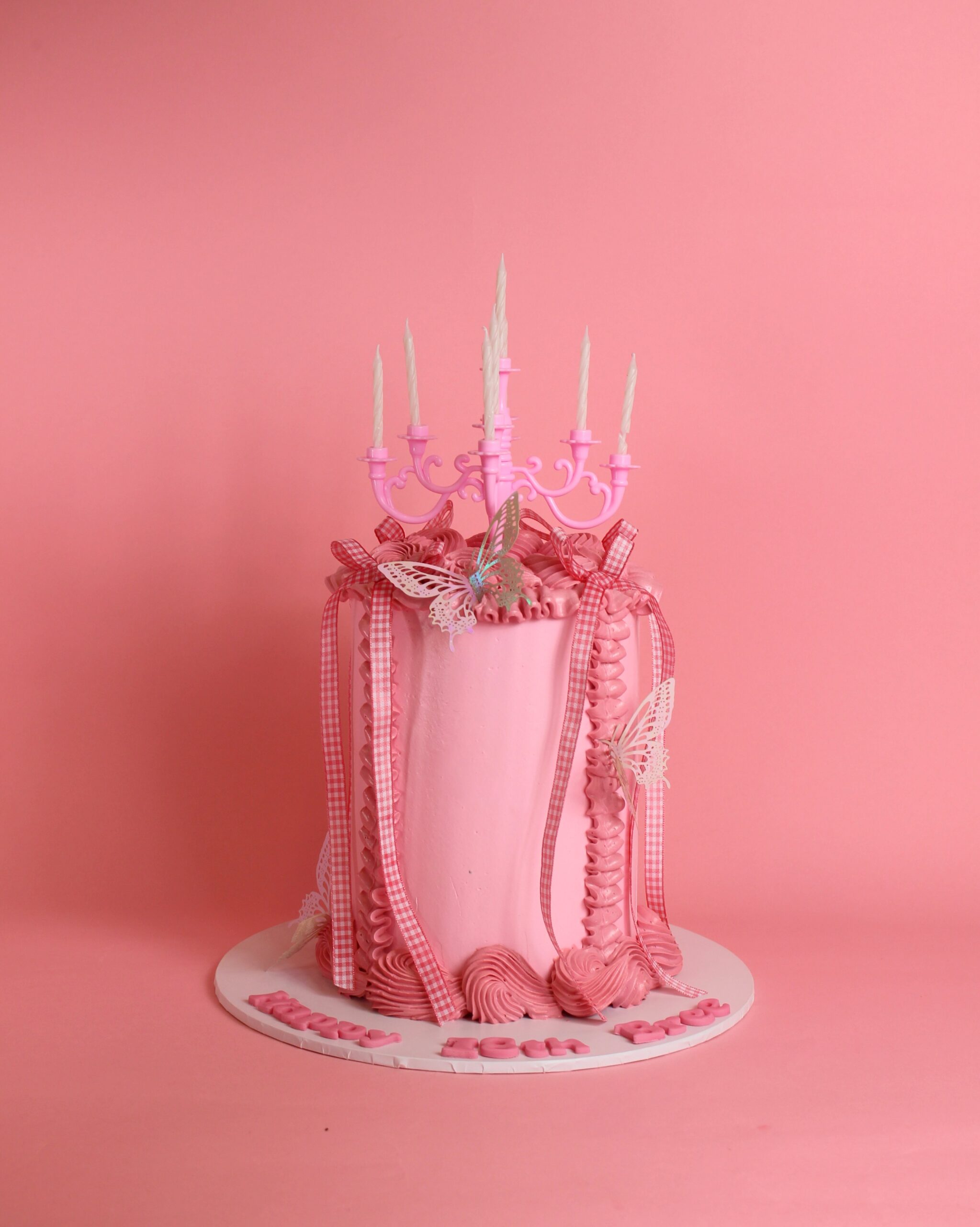 A pink birthday cake adorned with decorative candles and butterflies on a pink background.