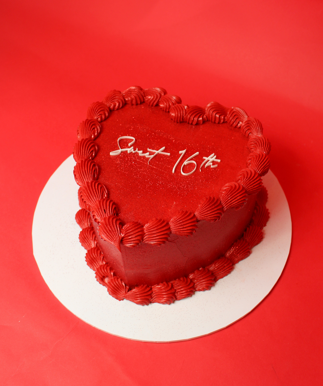 Heart-shaped red cake with red frosting and 'Sweet 16' written on top in white icing.