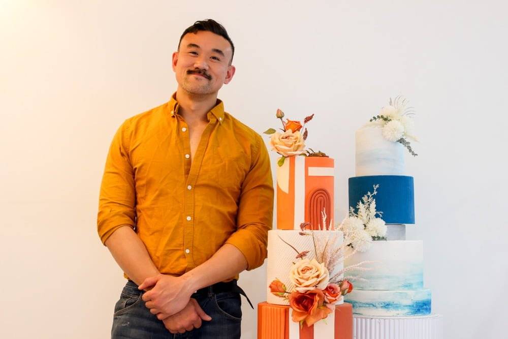 A man in an orange shirt standing beside three decorative cakes with floral designs.