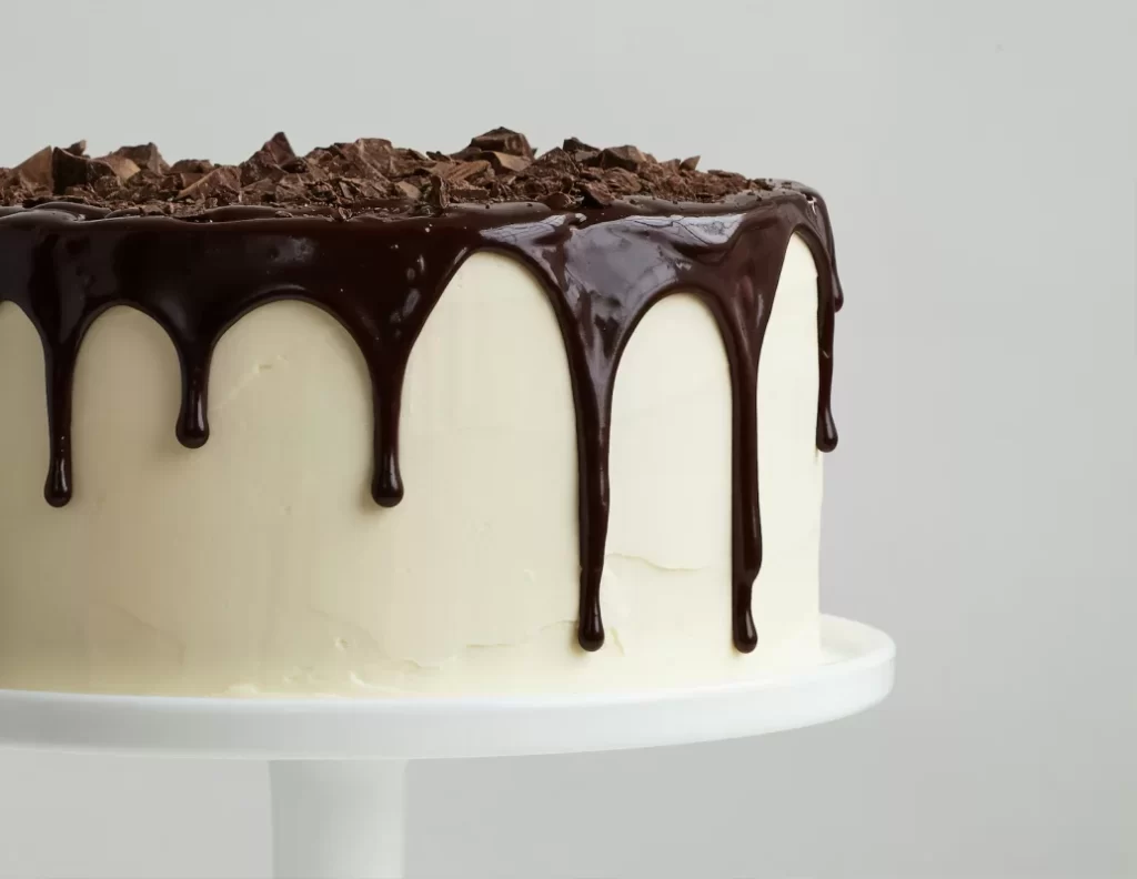A chocolate drip cake with a smooth white frosting and chocolate shavings on top.