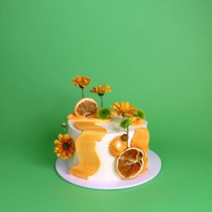A decorative cake adorned with orange frosting, dried orange slices, and yellow flowers against a green background. swirl piping cake, modern cake, comb piping , Squiggle cake, birthday cake