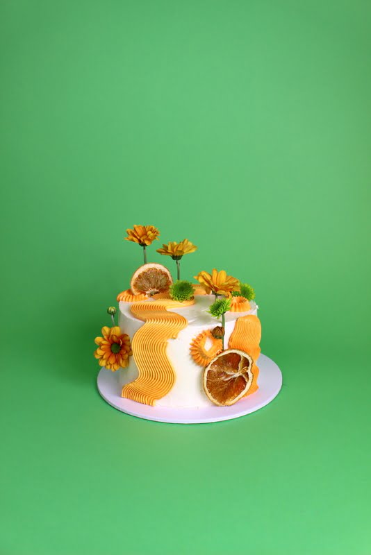 A decorative cake adorned with orange frosting, dried orange slices, and yellow flowers against a green background. swirl piping cake, modern cake, comb piping , Squiggle cake, birthday cake