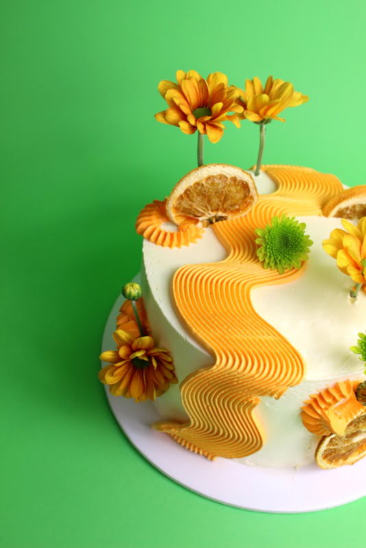 A decorative cake featuring orange and yellow icing, adorned with flowers and dried orange slices. swirl piping cake, modern cake, comb piping , Squiggle cake, birthday cake