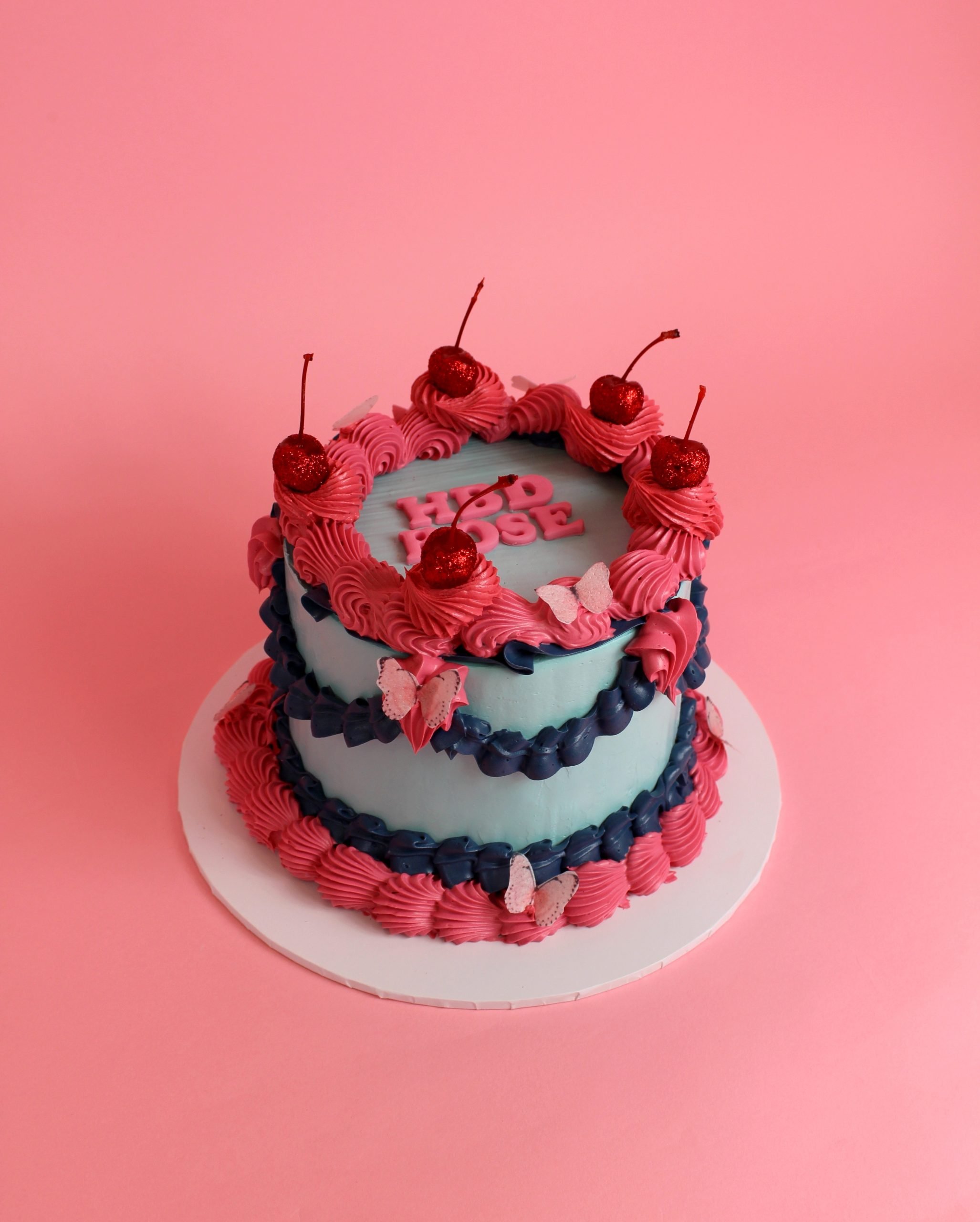 A two-tiered cake decorated with pink and blue frosting, topped with cherries and butterflies. vintage cake, retro cake, birthday cake