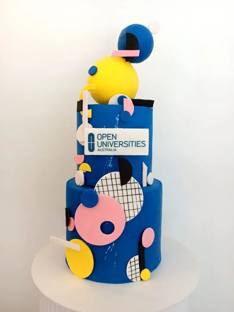 Two-tiered cake featuring the Open Universities Australia logo and colorful geometric decorations. corporate cake, business launch, company cake