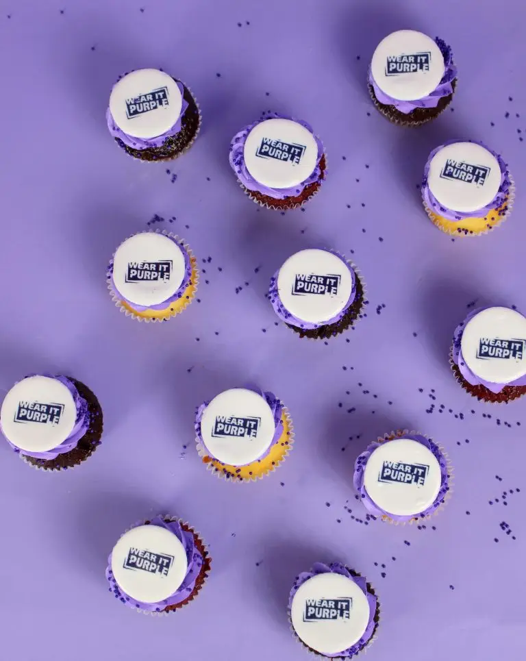 Cupcakes with purple frosting and white tops featuring a logo that says 'Wear It Purple'. corporate cake, business launch, company cake