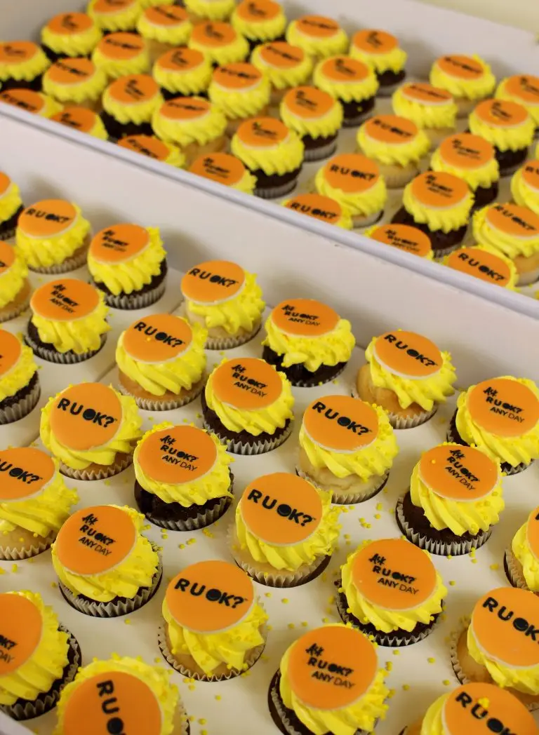 Tray of cupcakes with yellow frosting and orange RUOK? toppers arranged in a box. corporate cake, business launch, company cake