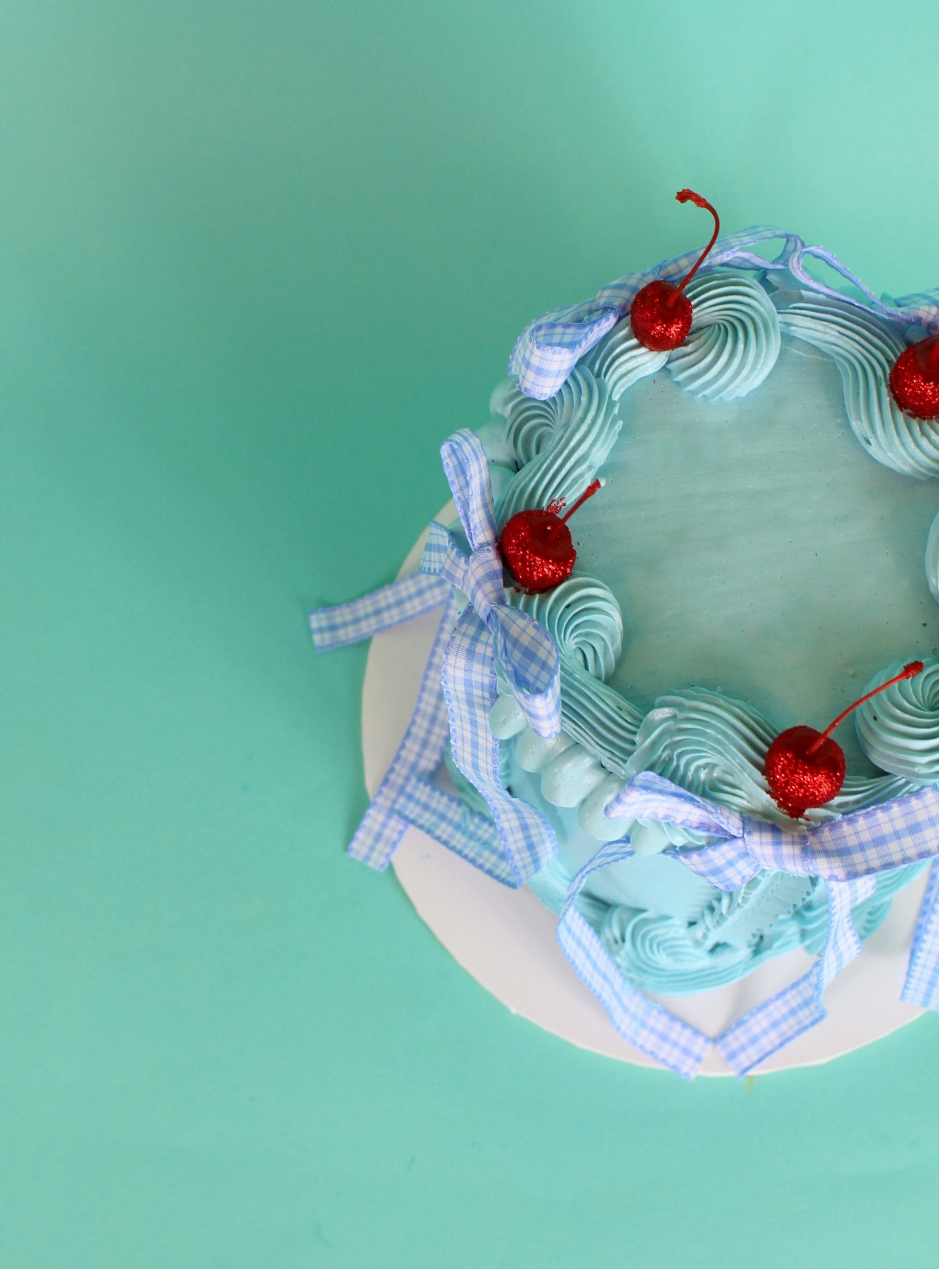 A blue frosted cake decorated with red cherries and blue ribbons on a white base. vintage cake, retro cake, dorothy cake, wizard of oz cake, birthday cake