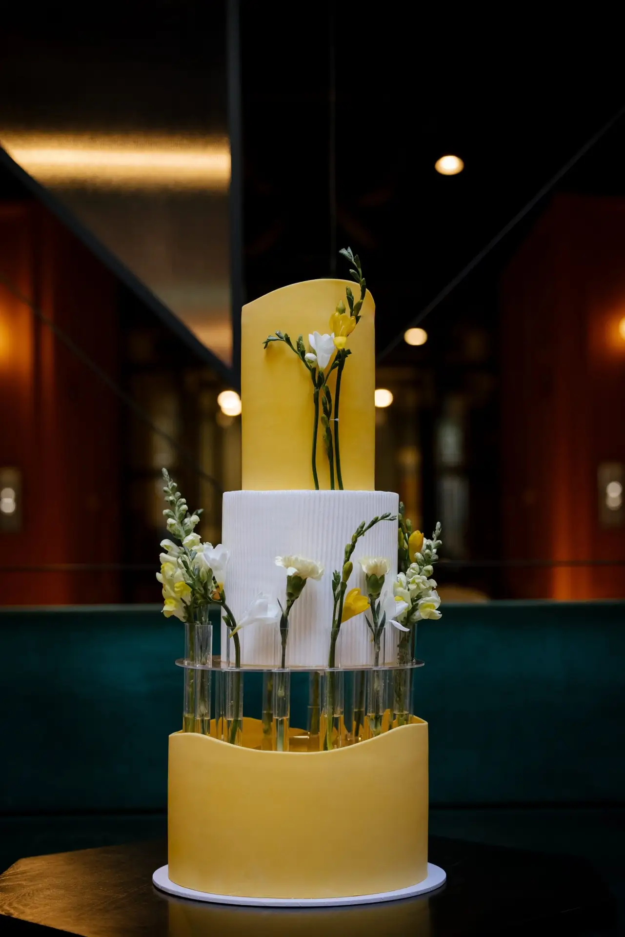 Three-tiered cake with yellow and white layers, adorned with flowers and placed in a glass container. wedding cake, floral cake, floral crown cake