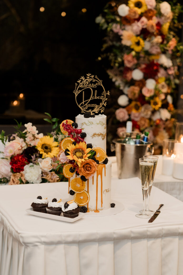 Two-tier white wedding cake with gold accents, orange slices, blackberries, sunflowers and roses.